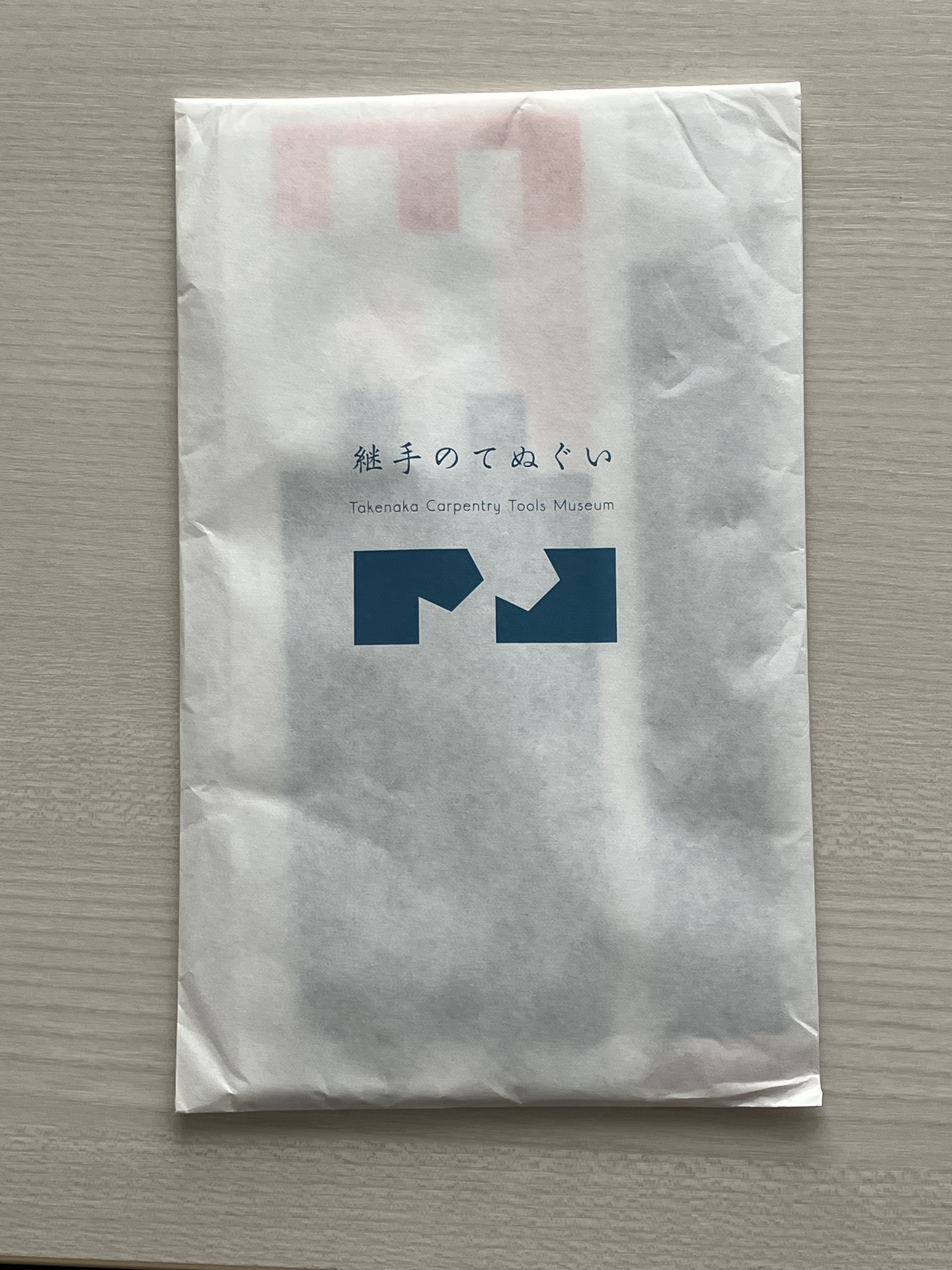 手ぬぐいが入っている紙袋。縦型で中央に継手のてぬぐいと書いてあり、その下にはTakenaka Carpentry Tools Museumと、その下には継ぎ手を模したロゴがある