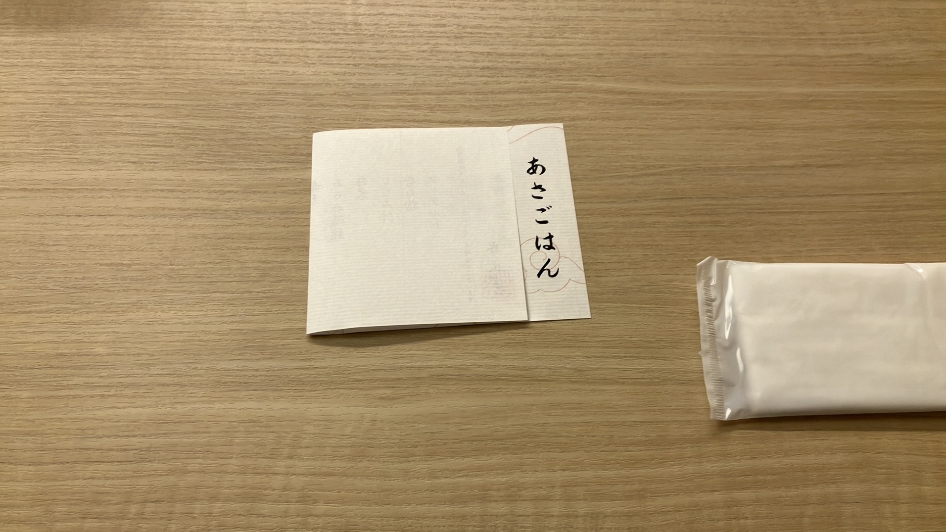 木目調のテーブル上に、中央には半分に折られてあさごはんと書かれた部分だけ見えるようになっているお品書きが、右側には見切れてお手拭きがある。